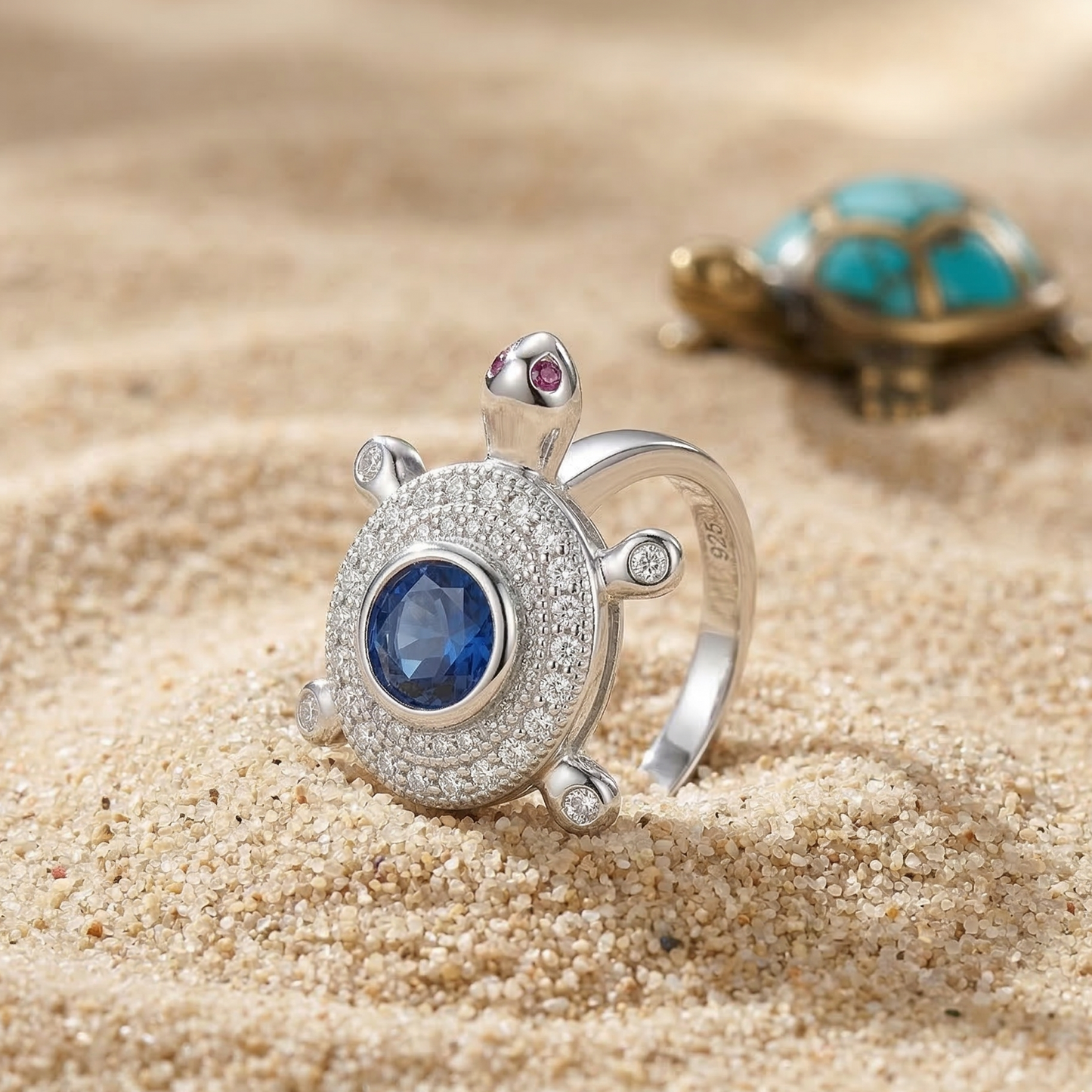 Silver turtle-shaped ring with a blue gemstone on sand, with a turtle figurine in the background.
